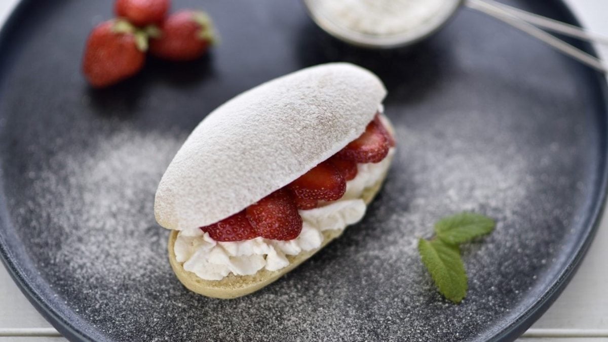 Auf einem Teller liegt ein veganer Brioche-Bun. Er is tmit Sahne und Erdbeeren gefüllt und mit Puderzucker bestreut.
