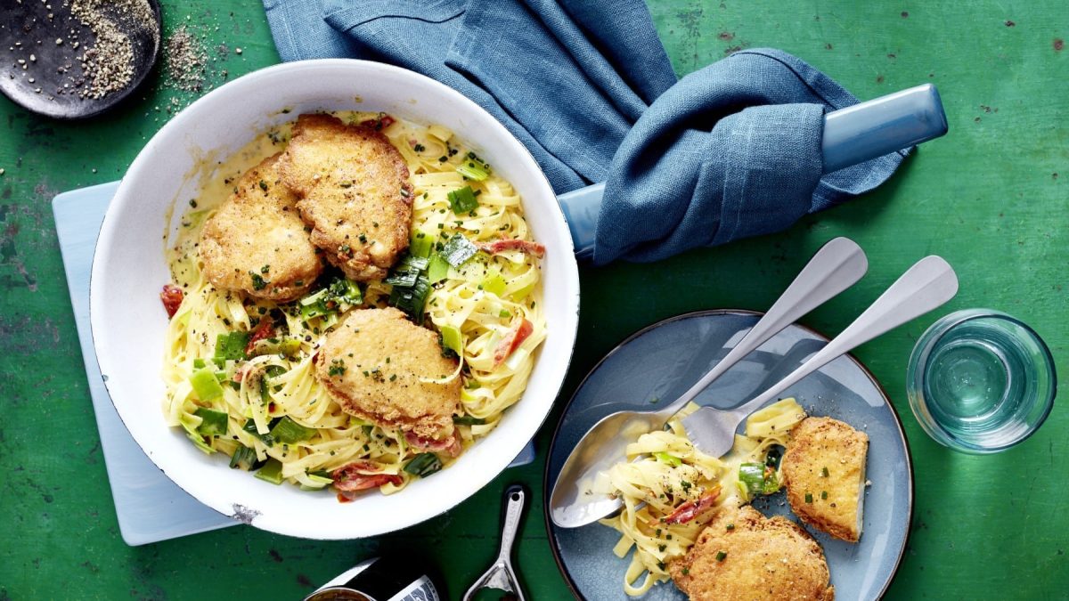 Parmesan-Schnitzel mit Porree-Carbonara, serviert in einer großen Schale und garniert mit Schnittlauch und grobem Pfeffer auf einem grünen Esstisch.