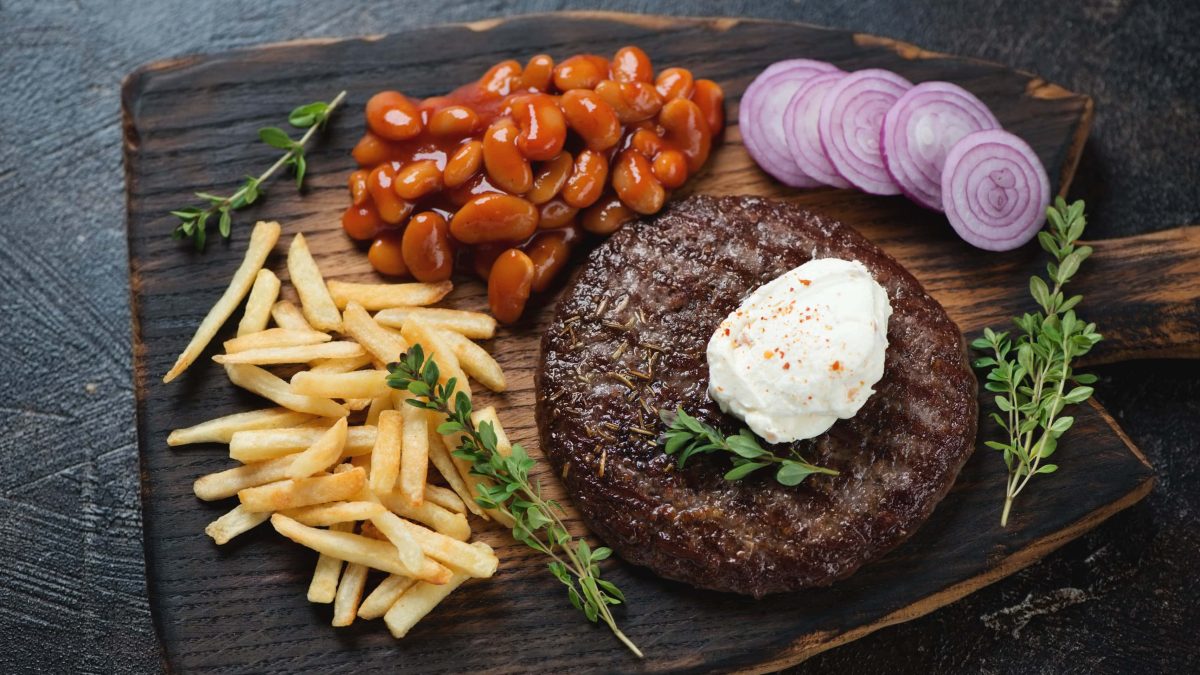 Pljeskavica mit Rahmkäse, Bohnen, Pommes, Zwiebeln und Kräutern auf Holzbrett. Draufsicht.