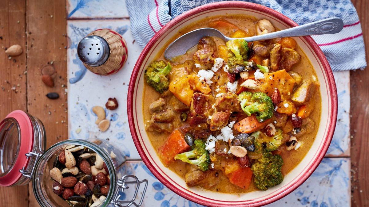 Putengulasch mit Kürbis und Honignüssen in einer Schüssel mit Löffel neben Salzstreuer und Nüssen vor hellem Hintergrund.