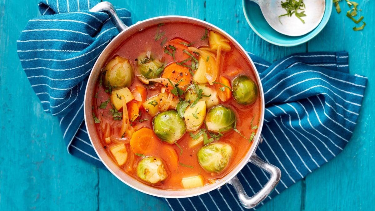 Rosenkohl-Kartoffel-Eintopf mit Möhren und frischen Kräutern, serviert im großen Topf auf einem blauen Tisch.