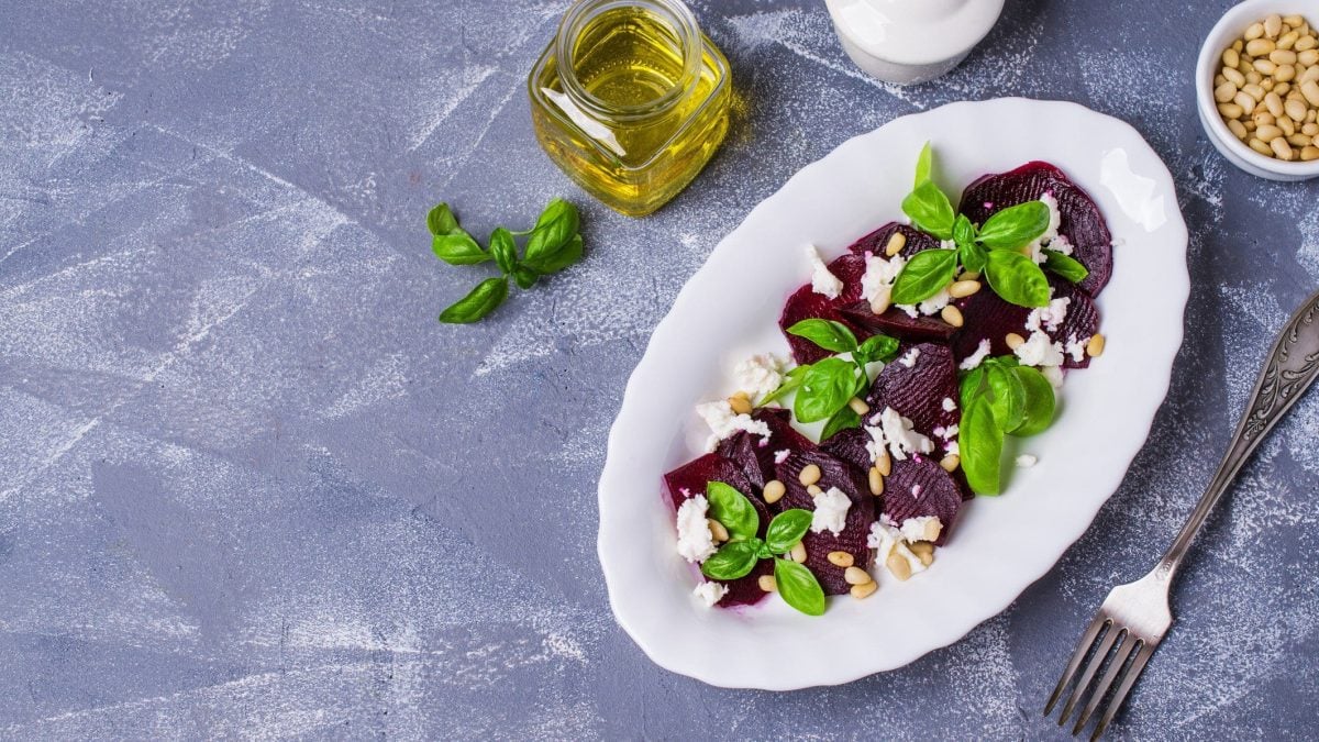 Rote-Bete-Carpaccio mit Feta auf blauem Untergrund, daneben Olivenöl und Pinienkerne