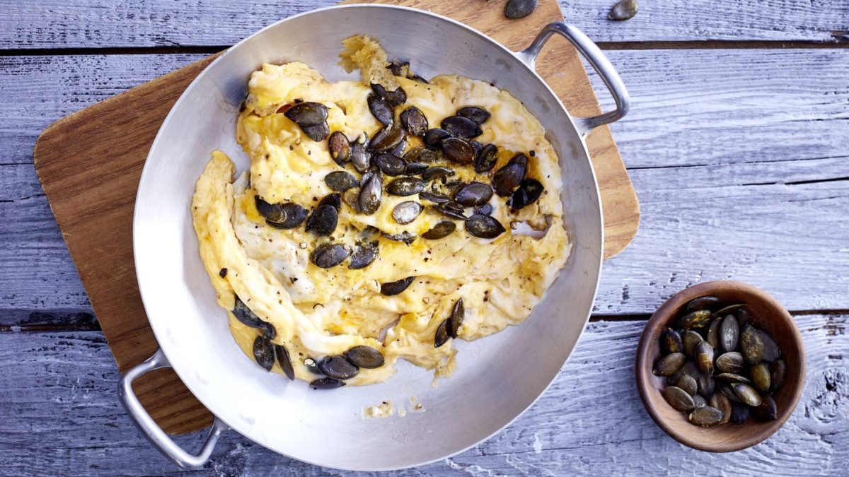 In einer großen Pfanne serviertes Rührei mit Kernen, garniert mit grobem Pfeffer und angerösteten Kürbiskernen, serviert auf einem hellen Holzbrett auf einfachem, grauem Holztisch.