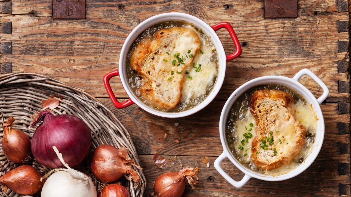 Soupe à l'ognion in zwei Schalen. Daneben ein Korb mit Zwiebeln und Knoblauch.