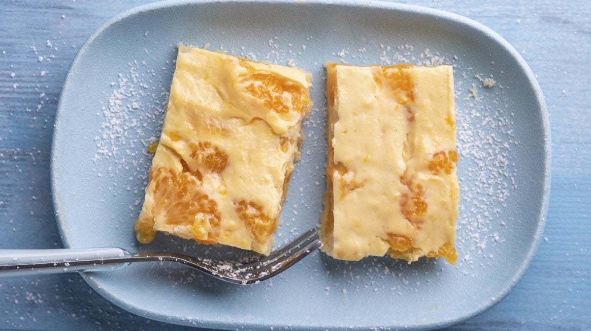 Auf einem Teller liegen zwei Stück veganer Mandarinen-Quark-Kuchen. Sie sind mit Puderzucker bestäubt.
