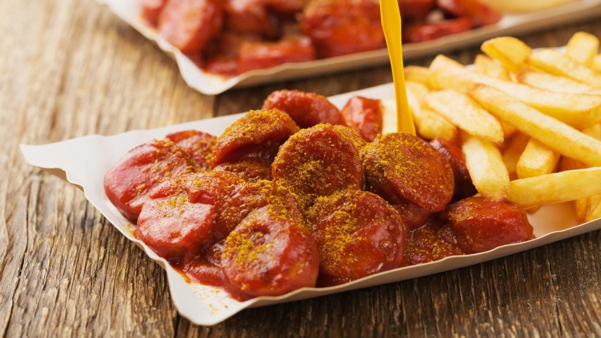 Original Berliner Currysauce mit Currywurst und Pommes und gelbem Plastikpiekser in Pappschale auf Holztisch. Im Hintergrunde eine weitere angedeutet. Frontalansicht.
