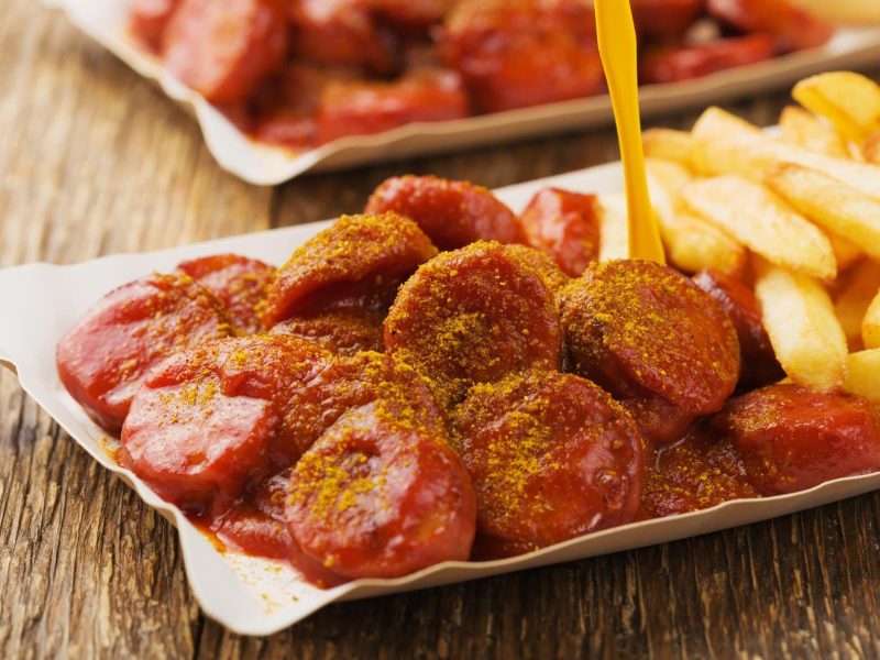 Original Berliner Currysauce mit Currywurst und Pommes und gelbem Plastikpiekser in Pappschale auf Holztisch. Im Hintergrunde eine weitere angedeutet. Frontalansicht.