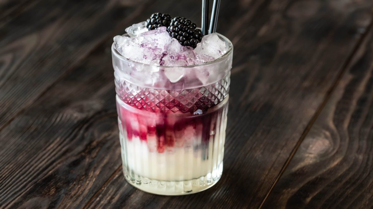 Bramble Gin-Cocktail mit Brombeeren in Tumbler mit frischen Brombeeren und Strohhalm auf dunklem Untergrund. Frontalaufnahme.