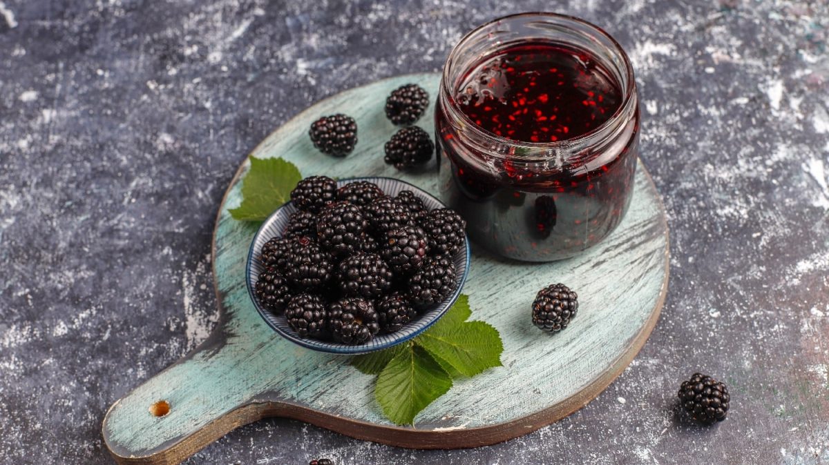 Ein Glas Brombeer-Marmelade auf einem Holzbrett mit frischen Brombeeren.