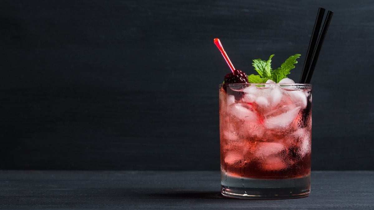 Ein rötlicher. durchsichtiger Drink mit Eiswürfeln, zwei Strohhälmen, Brombeere und Minze vor dunklem Hinter- und Untergrund.