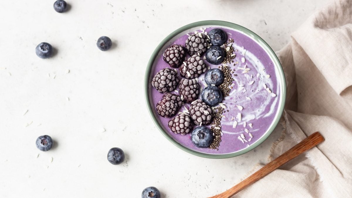 Eine Brombeer-Smoothie-Bowl mit Kokosflocken und gefrosteten Brom- und Heidelbeeren in der Draufsicht, daneben liegen ein paar Heidelbeeeren.
