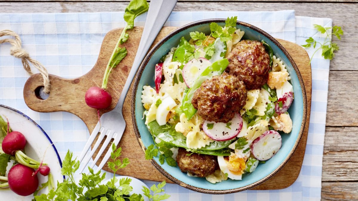 Buletten mit Nudelsalat in einem Teller auf einem Holzbrett neben Radieschen und einer Gabel.