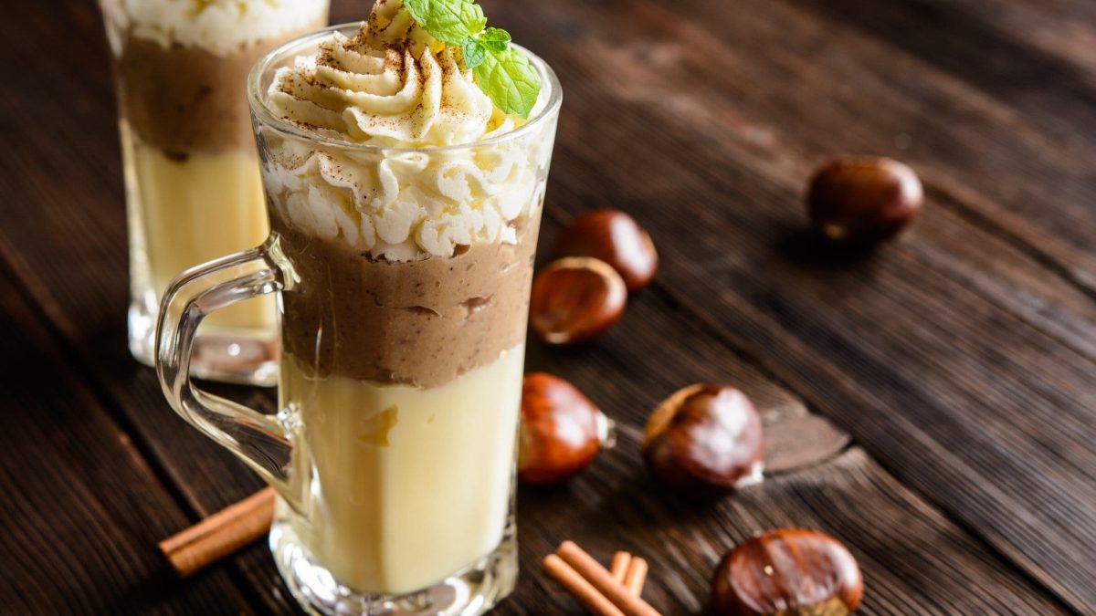 Cognac-Vanillepudding mit Maronenpüree und Pistazien-Sahne in Glas auf dunklem Holz. Daneben Maronen und Zimtstangen. Frontalansicht.