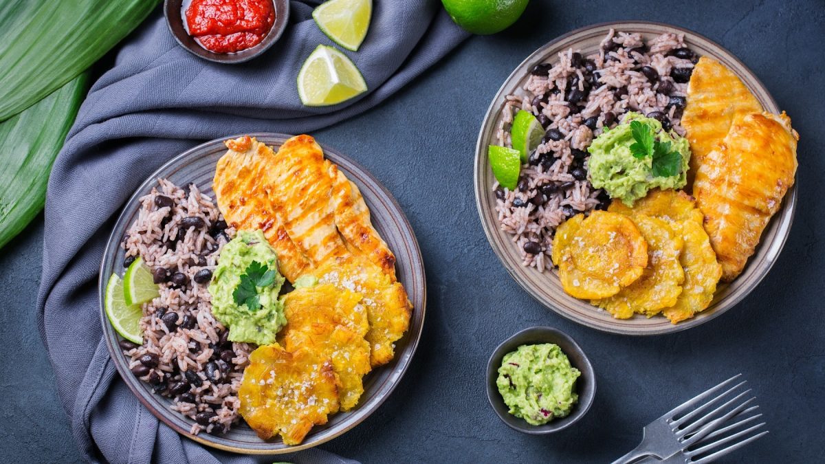 Zwei Teller mit Arroz Congrís con Tostones y Pollo neben kleinen Schalen mit Guacamole und Tomatensauce vor dunklem Hintergrund.