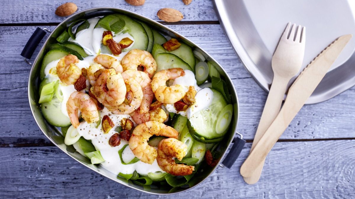 Curry-Garnelen mit Gurkensalat in einer Brotdose. Daneben liegt hölzernes Besteck.
