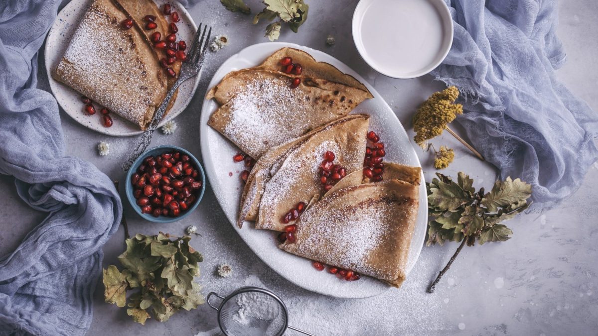 Einige Dinkel-Crêpes mit Granatapfelkernen auf Tellern neben herbstlicher Deko vor hellem Hintergrund.