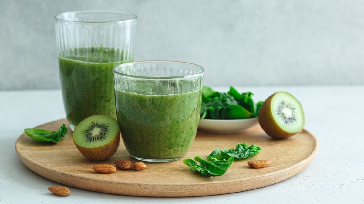 Zwei Gläser Spinat-Smoothie mit Kiwi auf einem Holzbrett neben frischen Früchten.