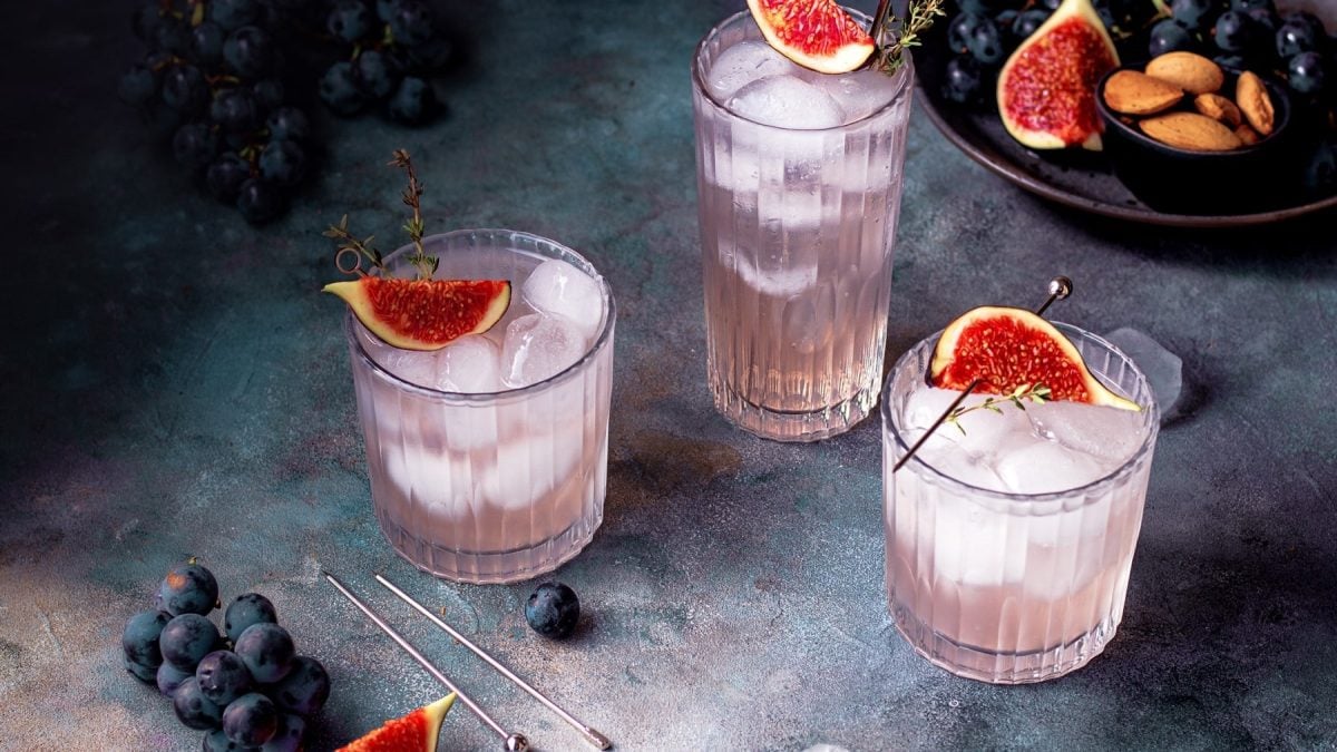 Drei Gläser mit Feigen-Fizz, Eiswürfeln, frischen Feigen und Thymian. Daneben Weintrauben und Mandeln.