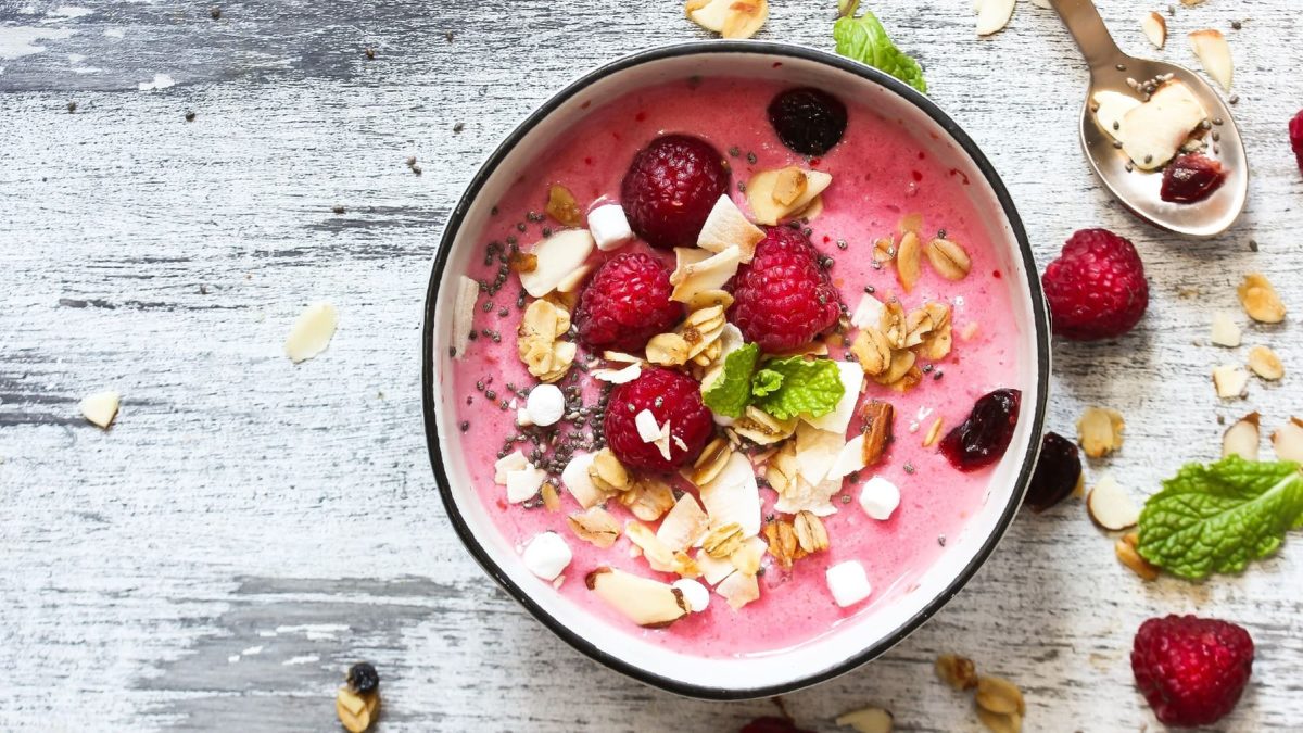 Eine Schüssel mit dem Himbeer-Kokos-Smoothie auf einem grauen Holztisch. Obendrauf frische Himbeeren, Kokoschips und Minze.