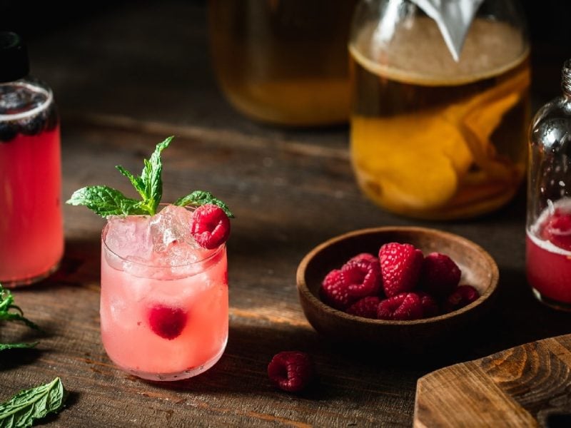 Ein Glas mit dem Himbeer-Kombucha auf einem dunklem Holzbrett mit allen weiteren Zutaten.