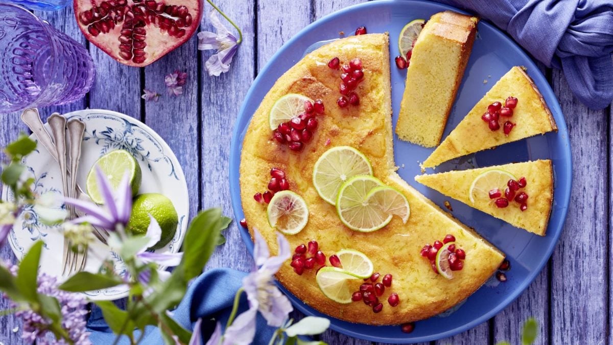 Angeschnittenet Joghurt-Limetten-Kuchen mit Granatapfelkernen und Limettenscheiben verziert auf einem blauem Teller neben Blumen und Geschirr.