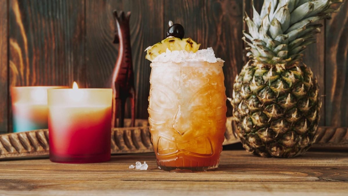 Jungle Bird Cocktail in Tikiglas auf Holz vor Holzwand. Im Hintergrund ganze Ananas, Kerzen und Holzdeko. Frontalaufnahme.