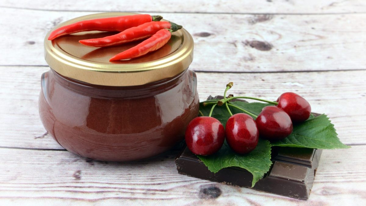 Chili-Kirschmarmelade mit Schokolade im Glas auf auf hellem Holz. Daneben Kirschen, Blätter und Schokolade. Frontalansicht.
