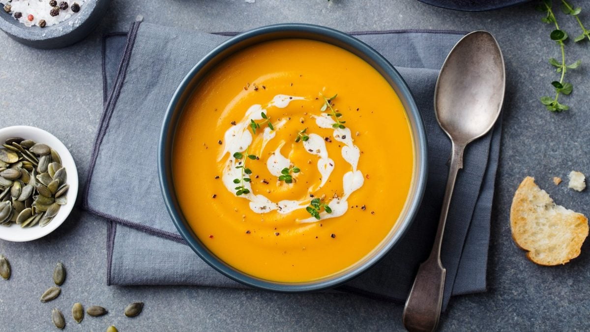 Eine Schale Kokos-Kürbissuppe in der Draufsicht, daneben ein Löffel und eine Scheibe Baguette sowie eine Schüssel mit Salz und Kernen.