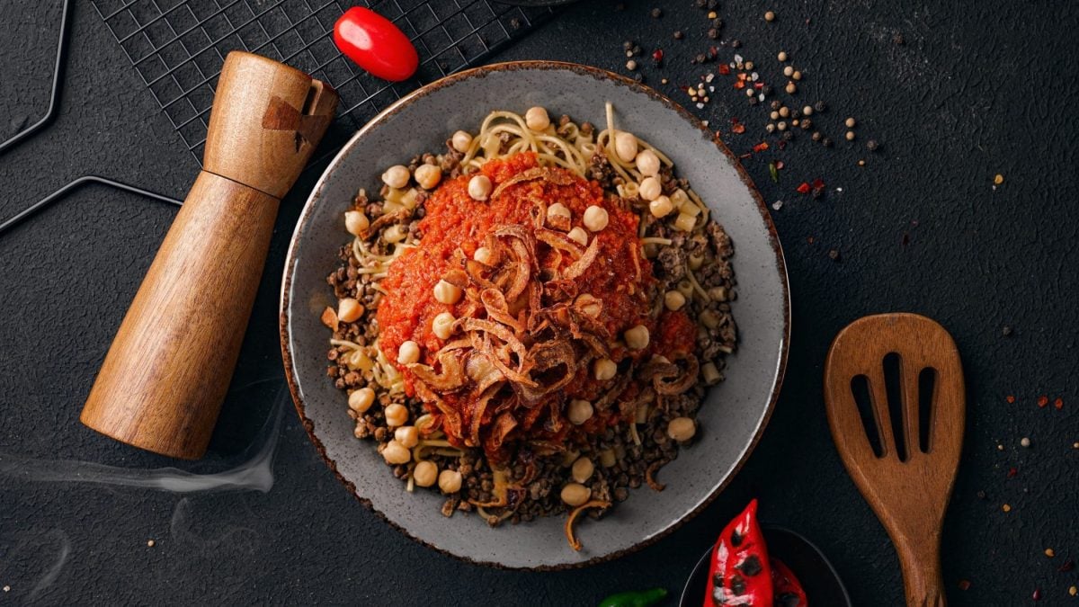 Koshari ägyptisches Streetfood auf grauem Teller und dunklem Untergrund. Danebem Pfeffermühle, Pfefferkörner und Küchenutensilien. Unten geröstete Chili. Draufsicht.