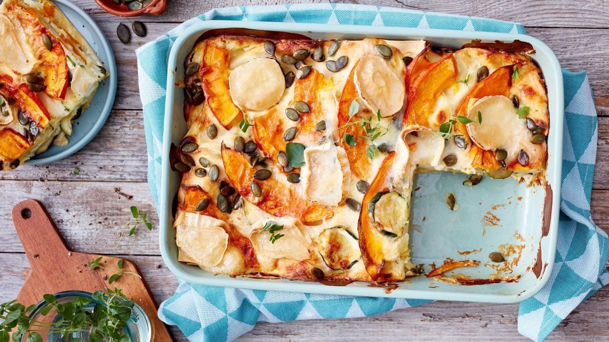 Kürbis-Ziegenkäse-Lasagne, bestreut mit Kürbiskernen und Schnittlauch, angeschnitten in einer Auflaufform serviert auf einem Holztisch.