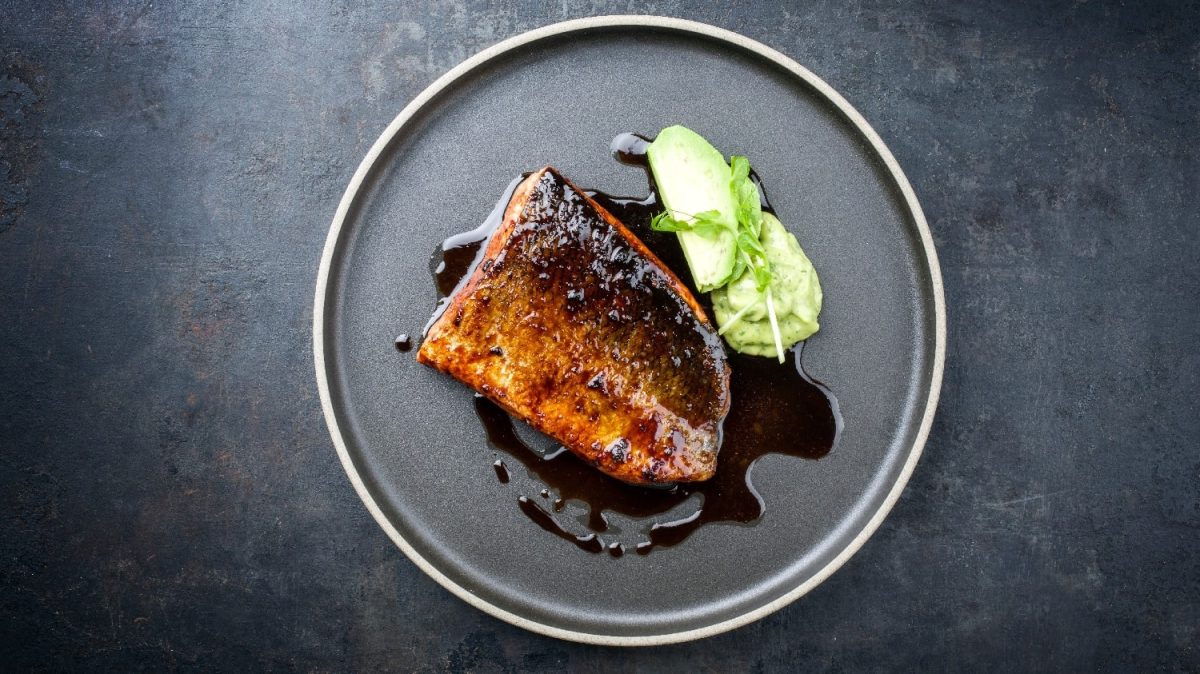 Ein schwarzer Teller mit dem Fishc und Sojalack. Rechts daneben ein Klecks mit der WAsabi-Kohlrabi-Creme und einem Stück Kohlrabi. Alles auf einem schwarzen Untergrund.
