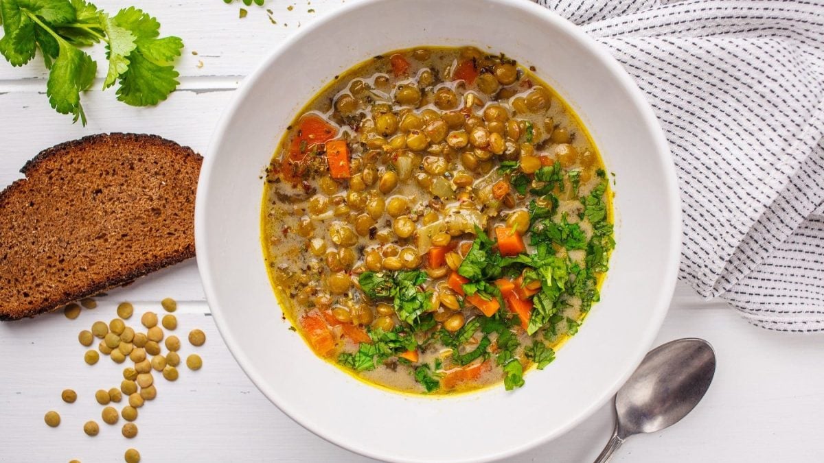 Aus der Vogelperspektive: Ein Teller Linsensuppe auf einem weißen Hintergrund. Drum herum liegen braune Linsen, eine Scheibe Brot und frische Petersilie