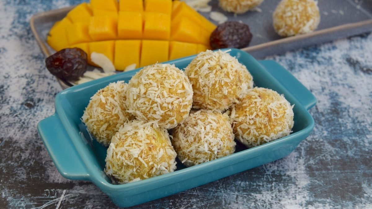 Ein paar Mango-Energy-Balls in einer blauen kleinen Auflaufform, dahinter eine angeschnittene Mango sowie Datteln.