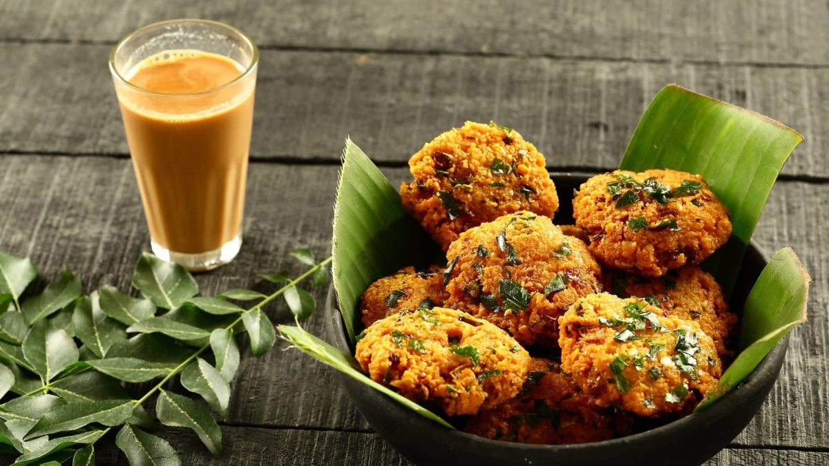 Masala Dal Vada in schwarzer Schale mit Blättern auf grauem Tisch. Links Curryblätter als Deko. Ein Glas mit Masala Chai oben im Bild. Draufsicht.