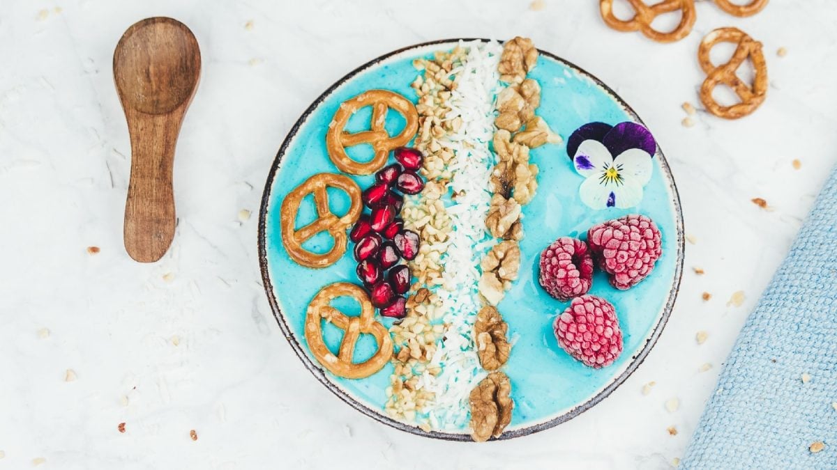 Eine Schüssel mit dem blauen Inhalt und den verschiedenen Toppings auf einem weißen Tisch. Daneben ein Holzlöffel udnd eine hellblaue Decke mit Brezeln.