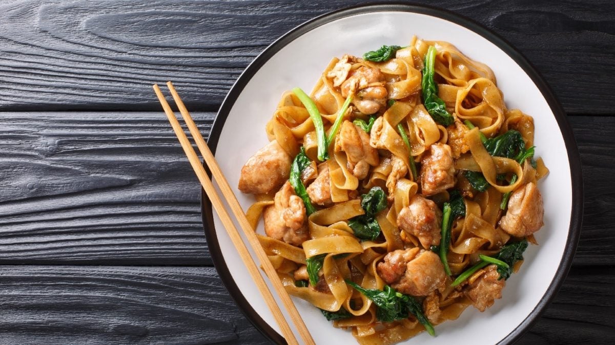 Gebratene Reisbandnudeln mit Hähnchen und Pak-Choi gebraten. Der Untergrund ist ein dunkler Holztisch.