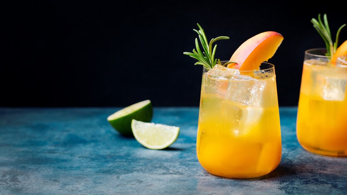 Zwei Gläser Pfirsich-Bourbon-Fizz mit Pfirsich und Rosmarin, daneben eine Limette.