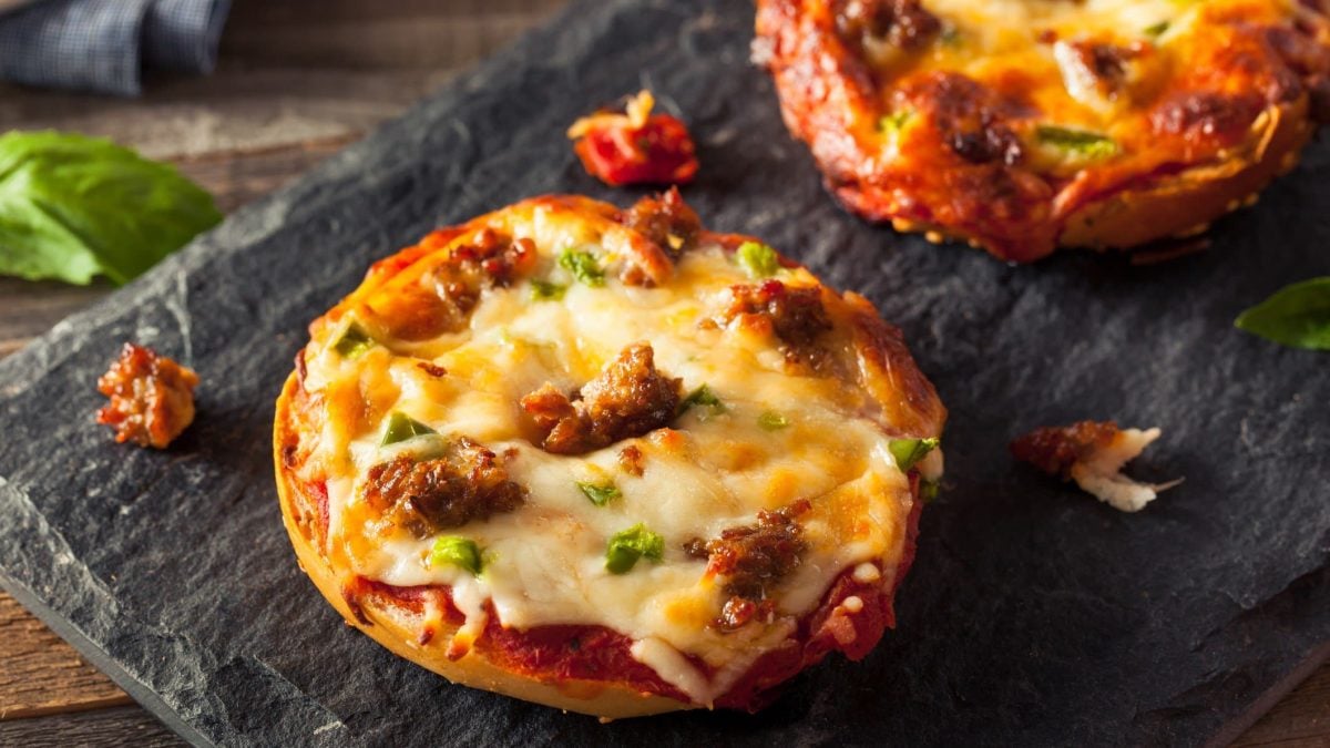Eine schwarze Schieferplatte, mit zwei Pizza Bagels auf einem Holzuntergrund. Daneben ein Zweig frischer Basilikum.