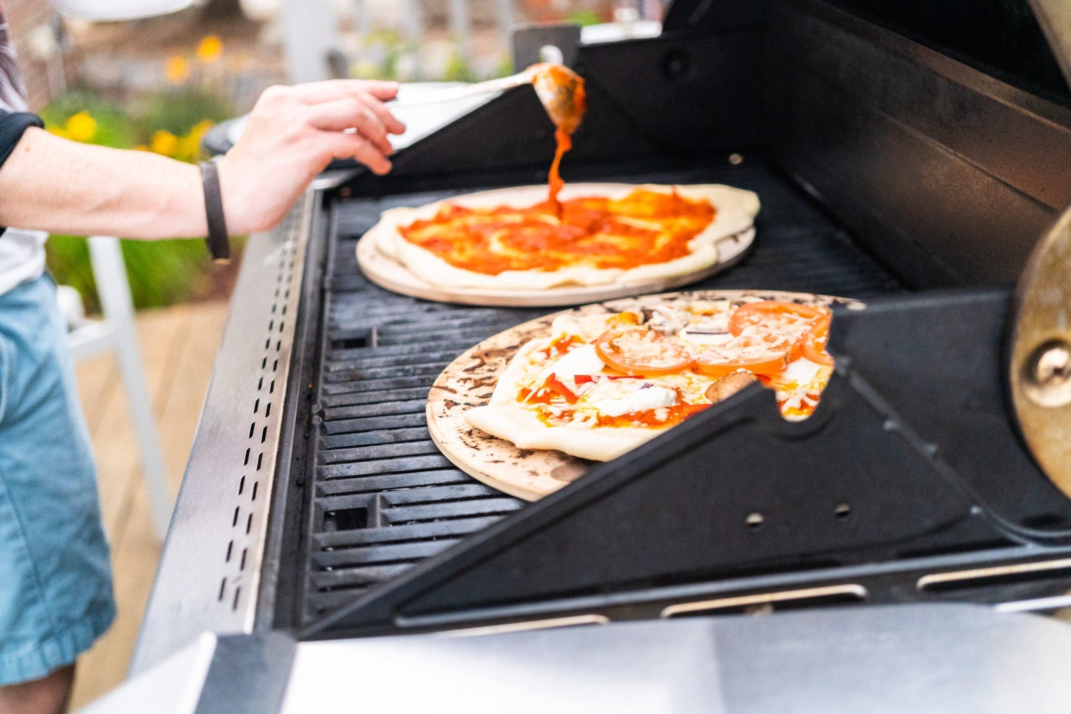 Zwei Pizzen auf Pizzastein auf Grill. Ein Mensch gibt Sauce auf die Pizza. Frontalansicht.