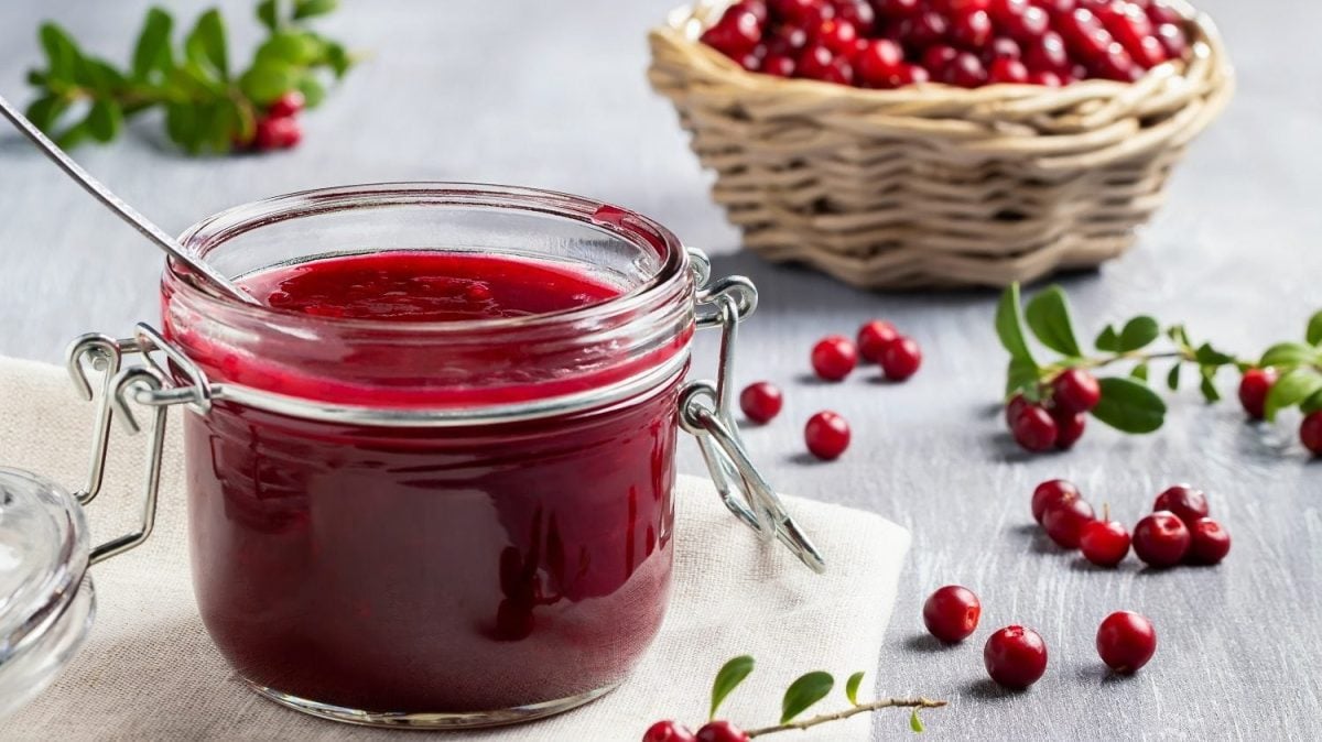 Frontalsicht: EIn Glas Preiselbeer-Konfitüre steht auf einem Tuch. drum herum liegen Preiselbeeren. Im Hintergrund steht ein Bastkorb mit frischen Preiselbeeren.