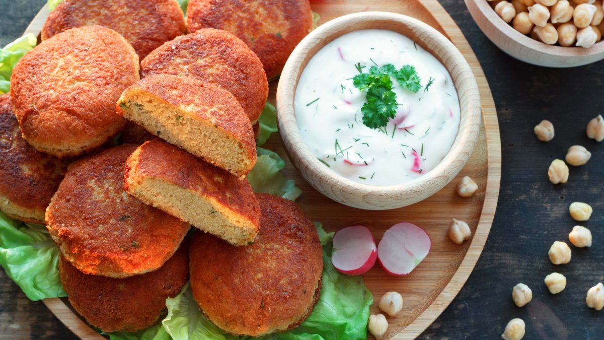 Radieschen-Raita mit Kichererbsenküchlein und Blattsalat auf Holzteller. Daneben Kichererbsen und Radieschen. Draufsicht.