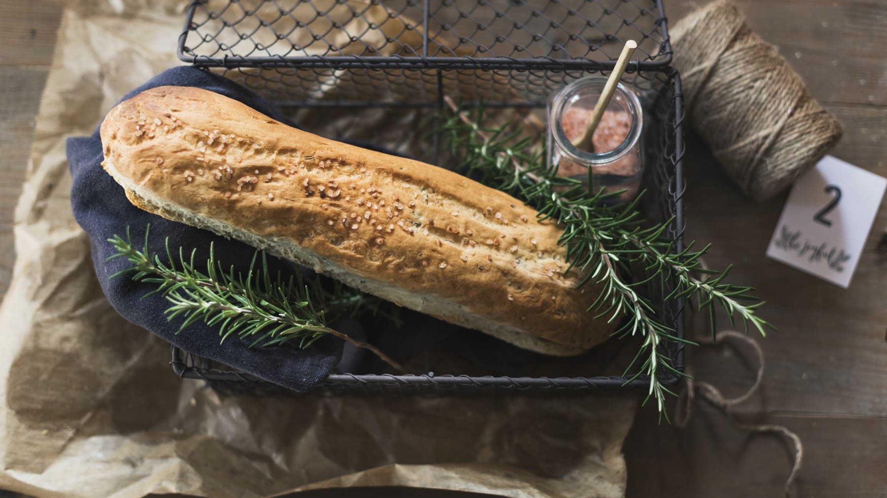 Rosmarin-Baguette dekoriert mit Rosmarinzweigen und rosa Steinsalz in einem schwarzen Drahtkorb vor braunem Hintergrund.