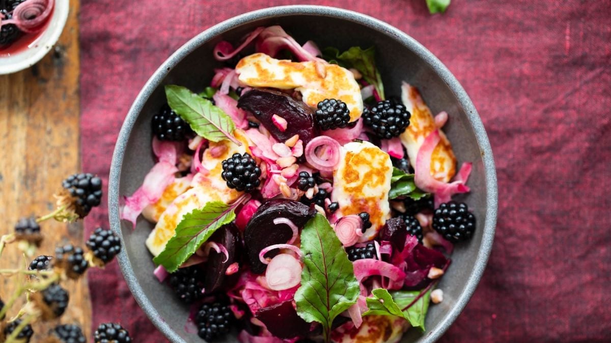 Eine graue Schale mit dem Halloumi-Salat auf einer weinroten Tischdecke. Unten links ist ein Stück vom alten Holztisch und ein Brombeerstrauch zu sehen.