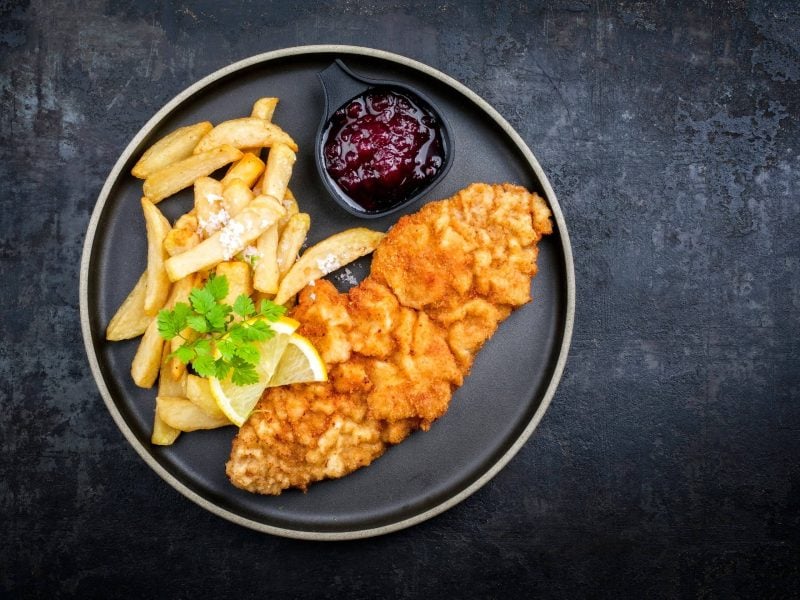 Ein dunkler Teller mit Schnitzel, Pommes, Preiselbeersauce und Zitrone auf einem dunklen Untergrund.
