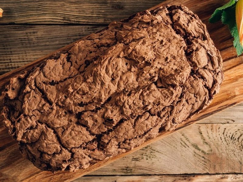 Draufsicht: Ein Kuchen auf einem Holzbrett.. Die Oberfläche ist nicht glatt, sondern rau. Es handelt sich um einen Zucchini-Schoko-Kuchen.
