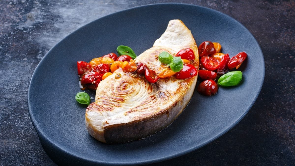 Ein schwarzer Teller mit dem Schwertfisch und Tomatenragout auf einem dunklen Untergrund.