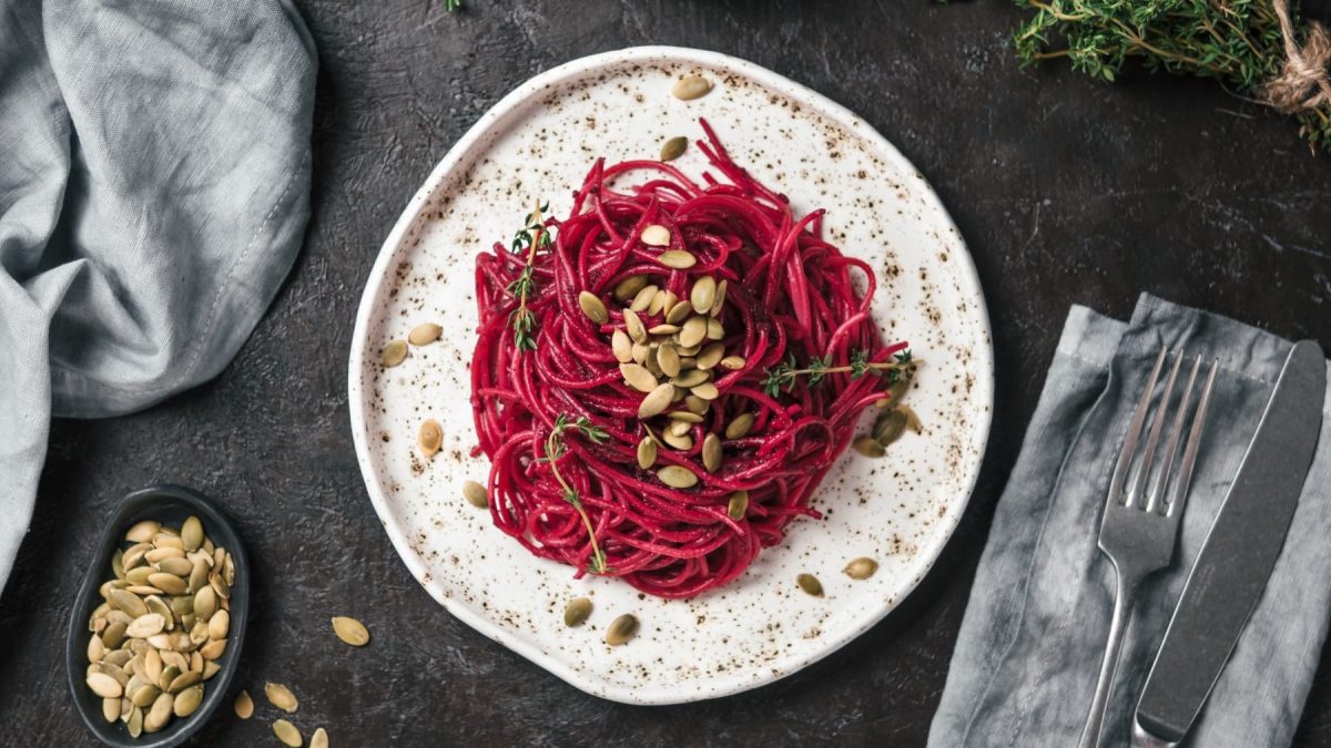 Spaghetti mit Rote-Bete-Pesto auf einem braunweiß gemusterten Teller. Daneben liegen verschiedene Zutaten sowie Besteck und Küchentücher.