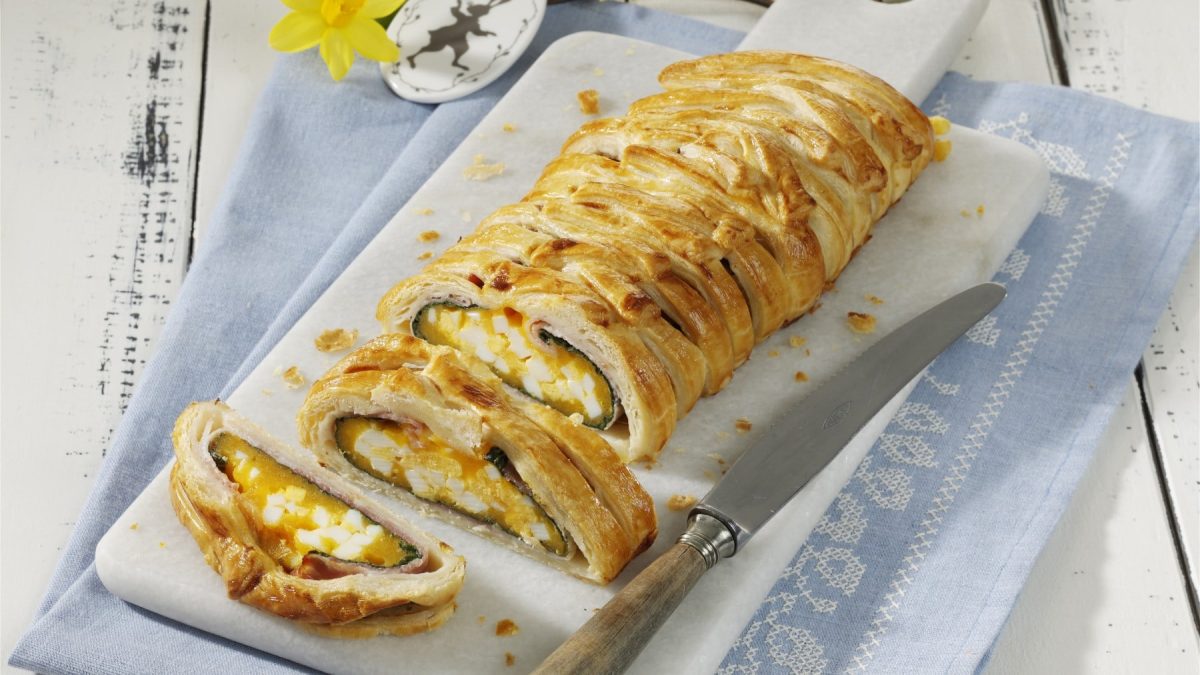 Angeschnittene Speck-Käse-Rolle auf weißem Schneidebrett vor hellem Hintergrund.