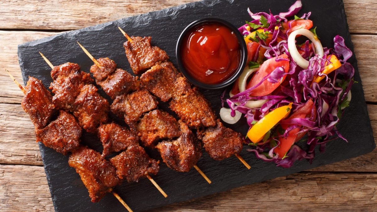 Suya Streetfood aus Westafrika mit Dipschale und Gemüsestreifen auf Schieferplatte. Draufsicht.