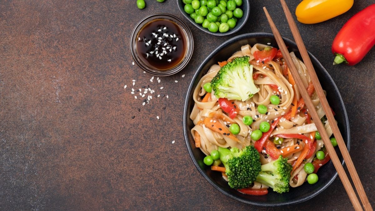 Ein dunkler Teller Teriyaki-Pasta mit Brokkoli, Paprika, Möhren und Erbsen, darauf Essstäbchen, daneben Gemüse, alles Draufsicht.